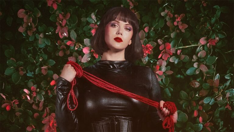 The beautiful Cincinnati, Ohio, professional dominatrix, Countess Cecilia, wearing a black latex bodysuit and corset holds shibari rope for a bondage session in front of a flower wall in her BDSM dungeon.