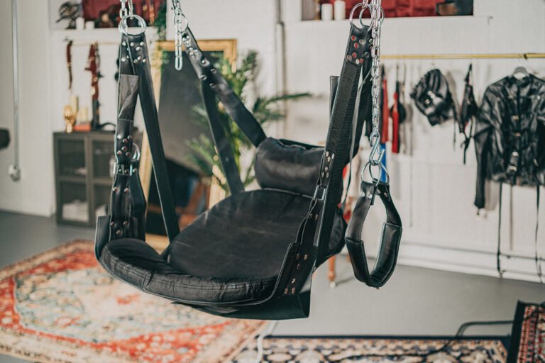 A leather sex swing hanging from the rafts of the Cincinnati Ohio BDSM dungeon of professional dominatrix, Countess Cecilia. In the background there is leather suspension suits, bondage jackets, and various floggers and whips.