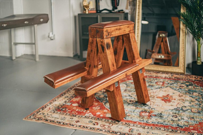 A custom built wooden spanking bench with leather padding used for impact play and corporal punishment sits in front of a mirror on a ornate rug in the Cincinnati Ohio BDSM dungeon of professional dominatrix, Countess Cecilia.