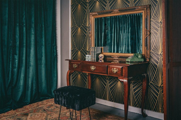 An elegant vanity area used for sissification with lush green curtains, art deco wallpaper, antique table, and a velvet stool awaits submissives in the Cincinnati Ohio BDSM dungeon of professional dominatrix, Countess Cecilia.