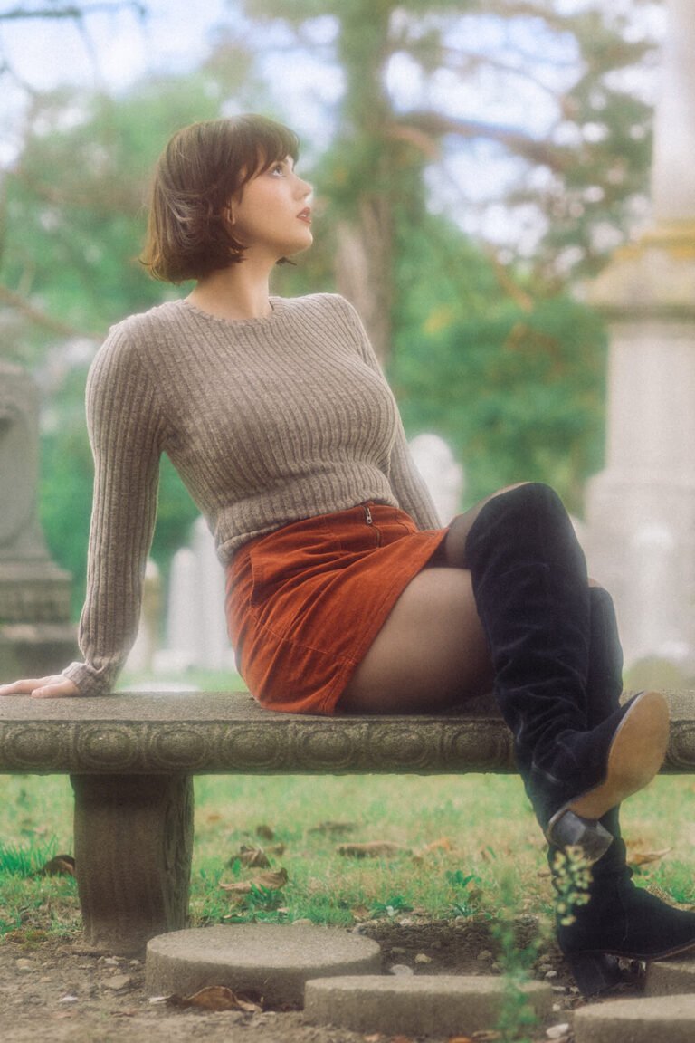 Cincinnati dominatrix and fetish model, Countess Cecilia, sits on a bench outside in a graveyard wearing a tight sweater, mini skirt, pantyhose, and knee-high black boots for this custom foot fetish content photoshoot.