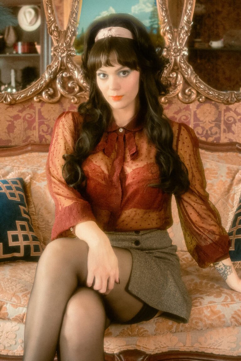 Cincinnati dominatrix and fetish model Countess Cecilia is dressed in a vintage wig, with a sheer red blouse, red lace bra, mini skirt, and pantyhose, while sitting on an antique couch in front of golden mirrors for this custom fetish content photoshoot.