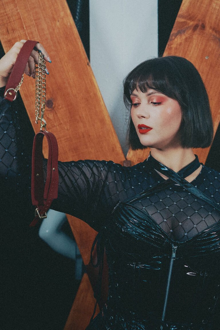 Cincinnati dominatrix and fetish model, Countess Cecilia, wearing a leather bodysuit, holds an empty leash in front of a St. Andrew's cross, waiting for a pet to wear in servitude.