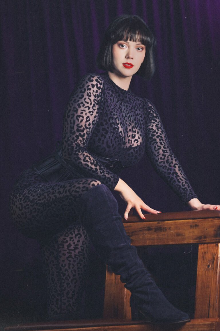 Cincinnati dominatrix Countess Cecilia in her dungeon, wearing a sheer leopard bodysuit with knee-high black boots, straddles a spanking bench used for BDSM and impact play.