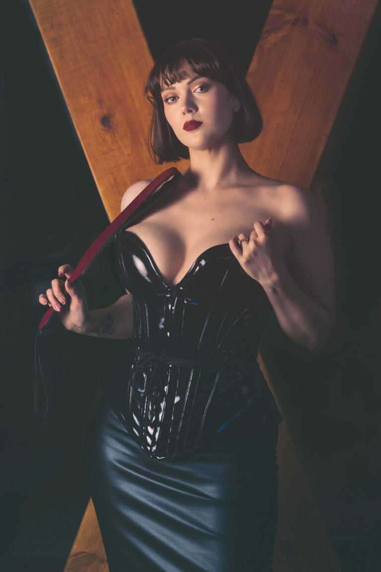 Cincinnati dominatrix Countess Cecilia wears a revealing leather corset, posing seductively in front of th St. Andrew's cross in her dungeon while holding a thick red flogger for her BDSM sessions.